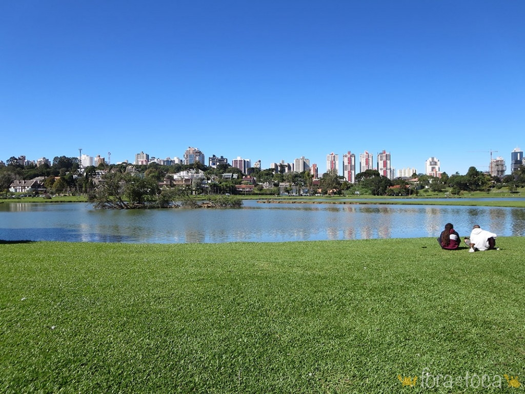 2 people sitting on the edge of the Lake of the Barigui Park with the buildings of the city in the background